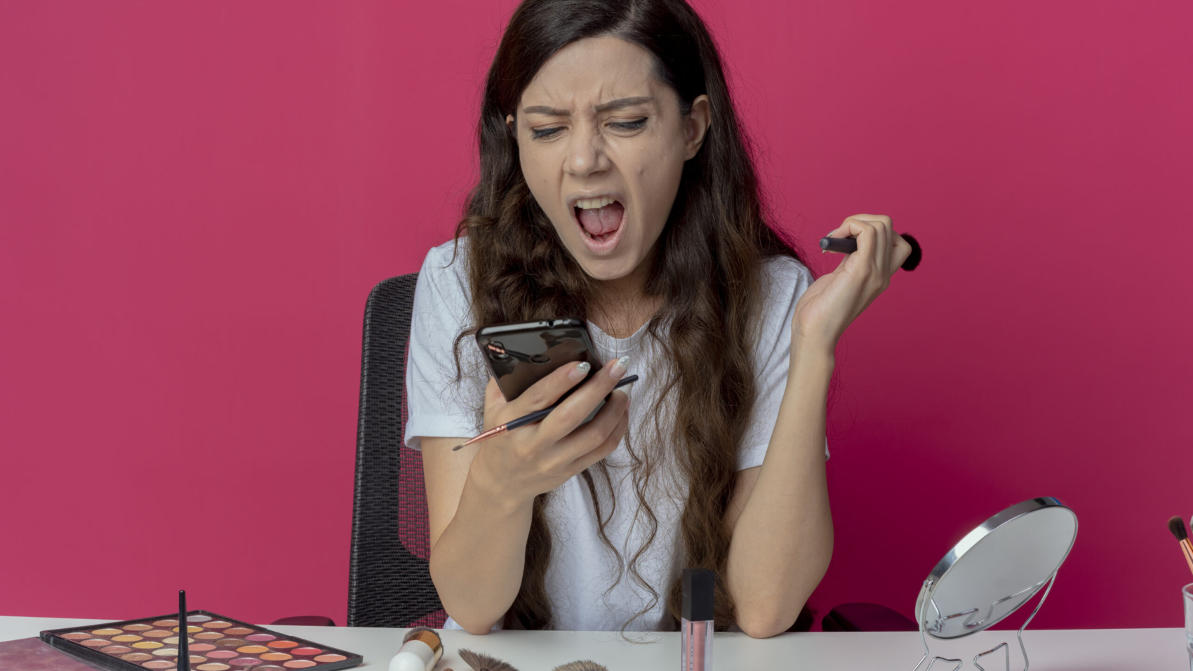 angry young pretty girl sitting at makeup table with makeup tools holding and looking at mobile phone and screaming with powder brush in another hand isolated on crimson background