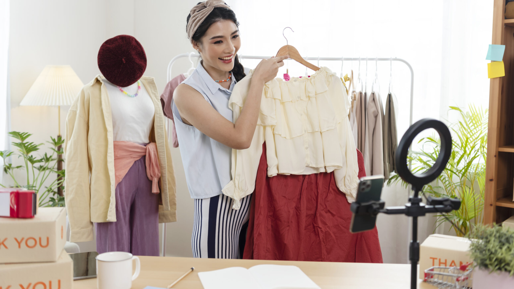 Asian young woman blogger showing clothes in front of smartphone camera while recording vlog video and live streaming at her shop. SME or freelance business online and delivery concept.