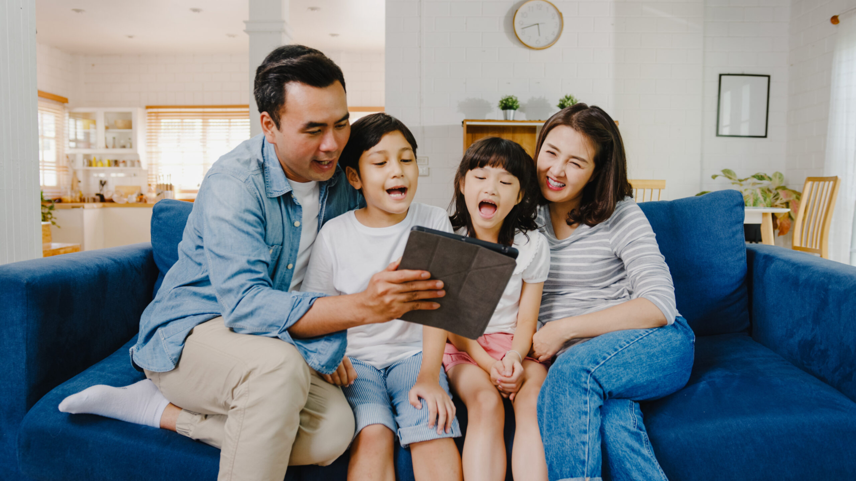 Happy cheerful Asian family dad, mom and kids having fun and using digital tablet video call on sofa at house. Self-isolation, stay at home, social distancing, quarantine for coronavirus prevention.