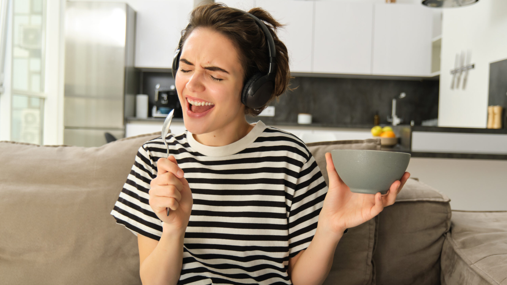 Young brunette woman holding bowl of cereals, singing with spoon in hand, listening to music in headphones, eating breakfast and enjoying favourite song, sitting on sofa at home.