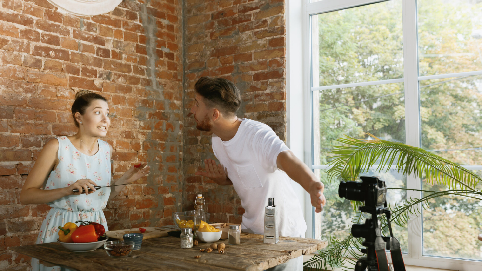 Vlogger and blogger freelance job, food concept. Young caucasian couple cooking together and recording live video for vlog and social media with professional camera against brick wall in their kitchen.